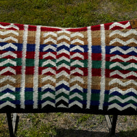 A contoured saddle blanket on saddle rack with white vertical lines and broken horizontal lines over a background of red, tan, gold, royal blue, and kelly green