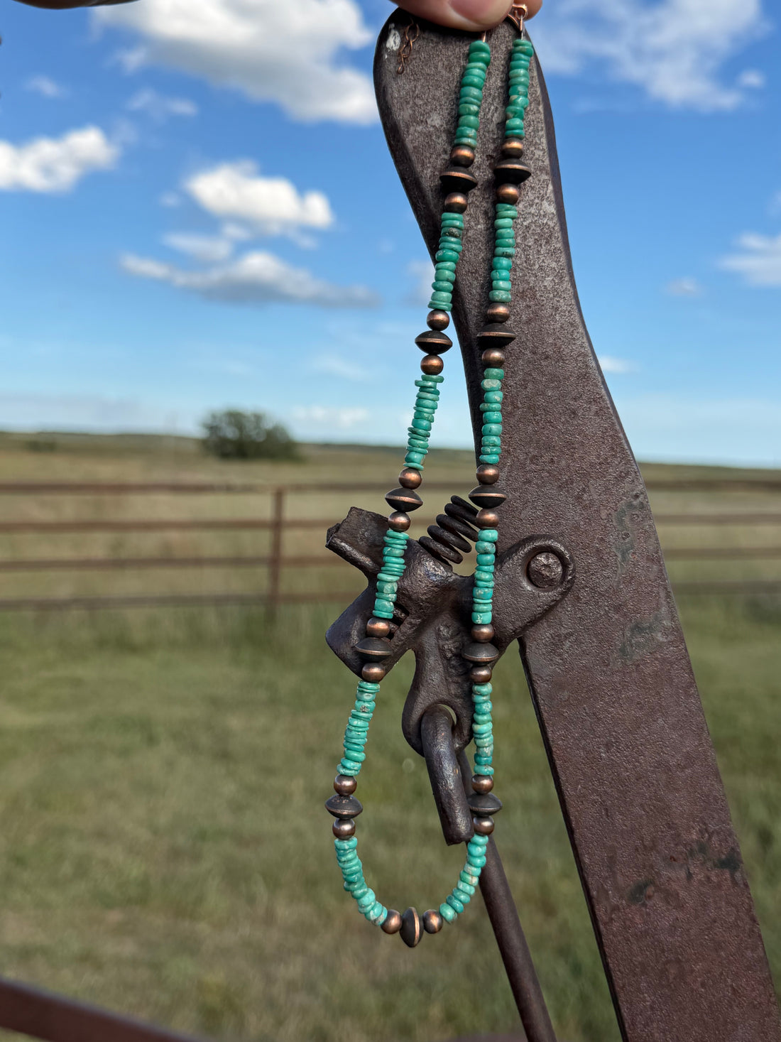 A turquoise and copper beaded necklace with a central charm, held up outdoors against a rusty metal gate with a grassy field and blue sky in the background.