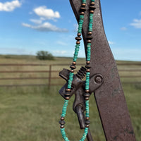 A turquoise and copper beaded necklace with a central charm, held up outdoors against a rusty metal gate with a grassy field and blue sky in the background.