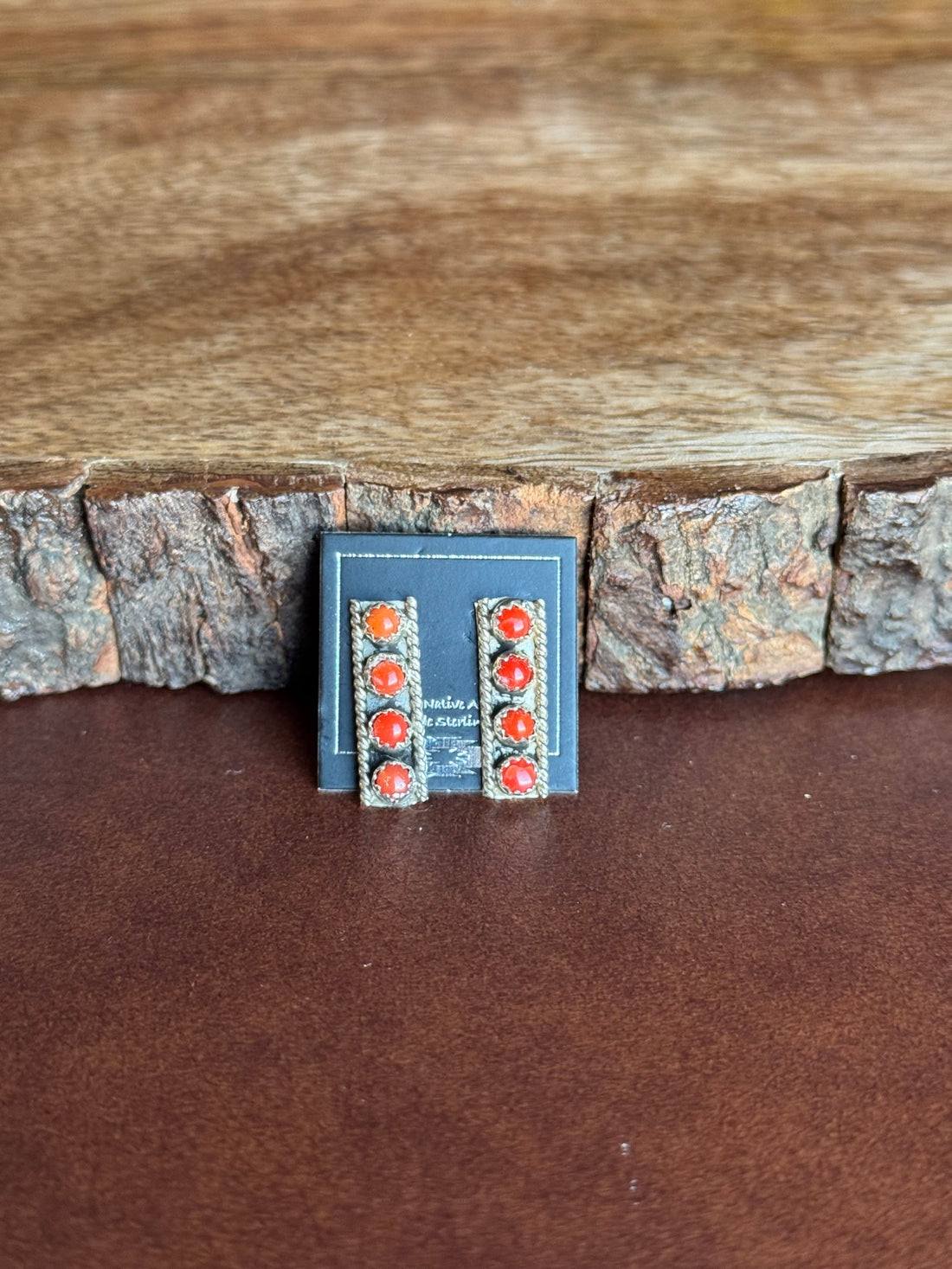 Silver vertical bar earrings set with red stones on a black card indicating native made leaning against wooden surface