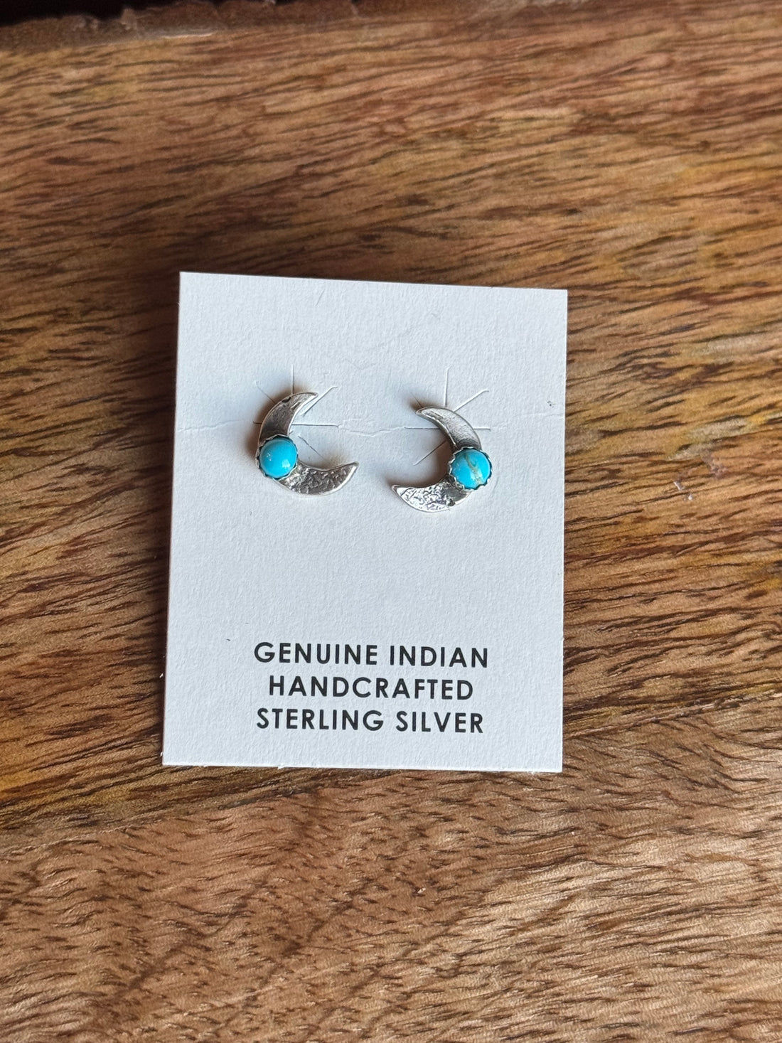 Pair of crescent moon silver earrings with turquoise stones on a card labeled 'Genuine Indian Handcrafted Sterling Silver' on a wooden surface.