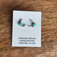 Pair of crescent moon silver earrings with turquoise stones on a card labeled 'Genuine Indian Handcrafted Sterling Silver' on a wooden surface.