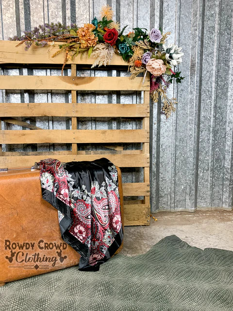 A collection of scarves with floral and paisley patterns draped over a suitcase, featuring colors such as black, red, white, and hints of yellow and pink.
