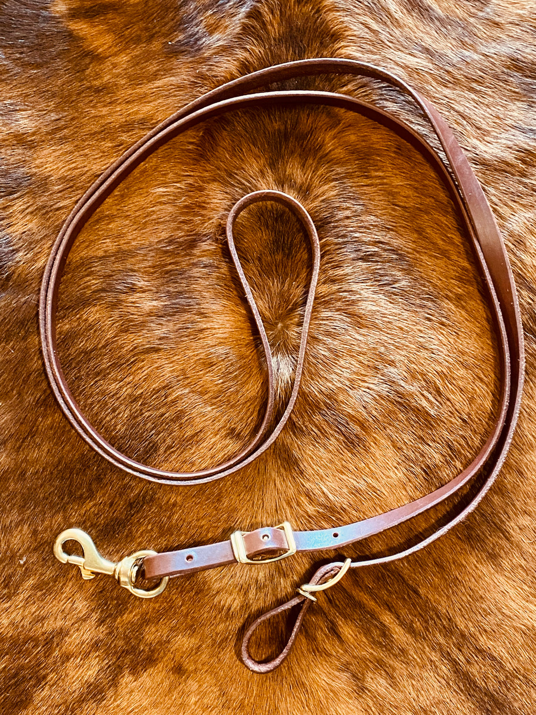 A brown leather rope rein laid out on a cowhide surface with brass buckles.